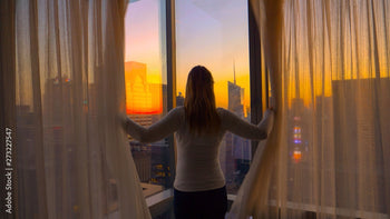 Person standing by a large window with a cityscape view during sunset.